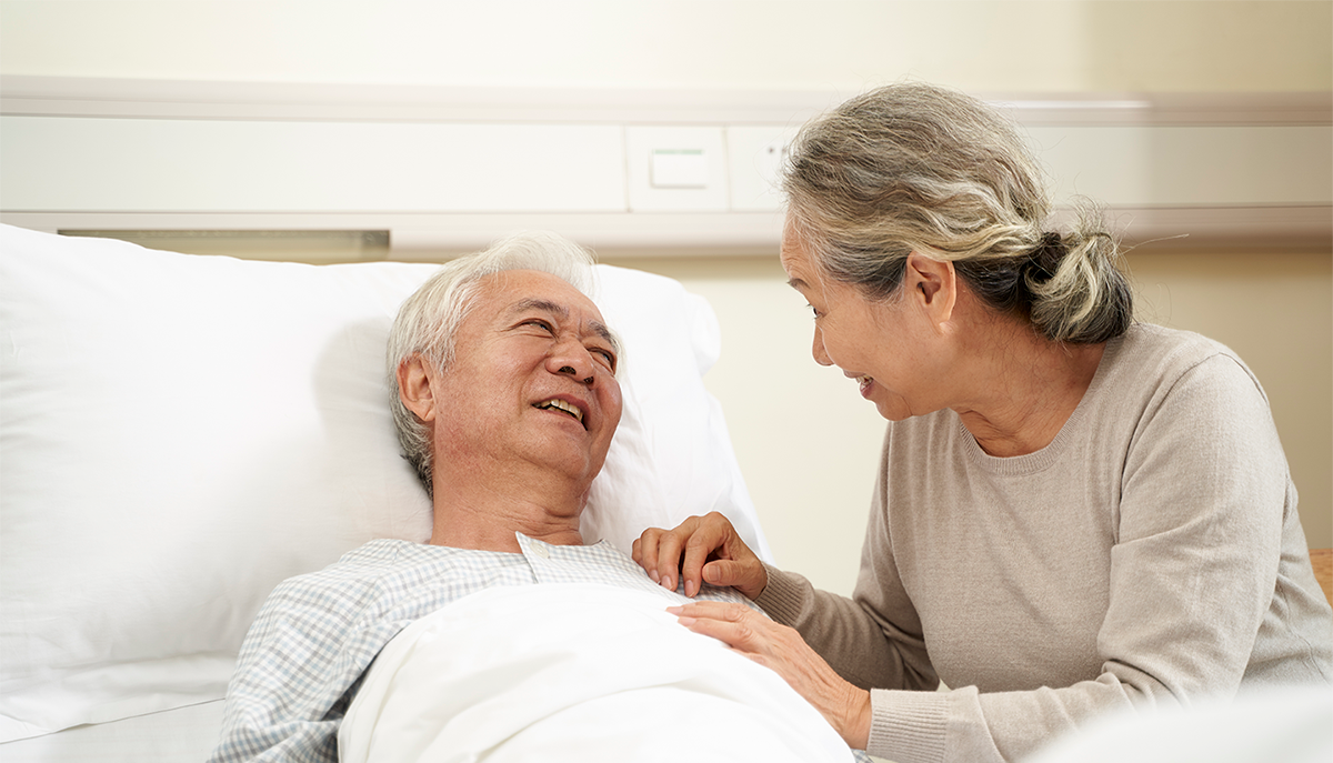 an elderly East Asian couple holding hands where one is in a hospital bed
